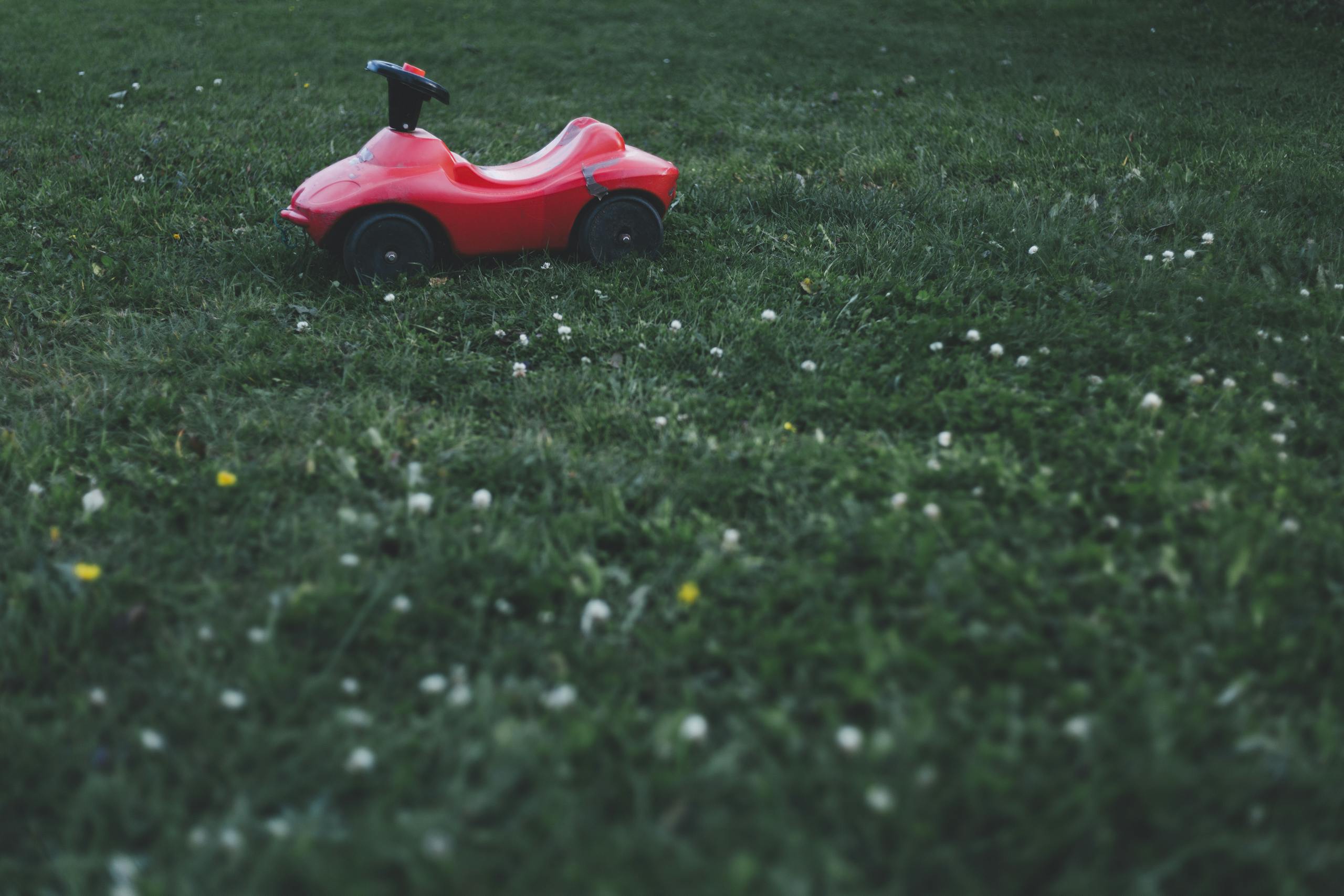 Red ride-on toy car sits on a lush green lawn, surrounded by tiny wildflowers.
